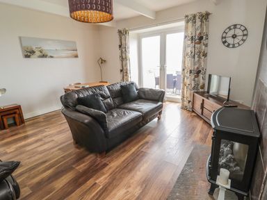 A living room with a black leather sofa a wooden TV stand with a TV and glass doors with patterned curtains at Seaview Howick in Craster