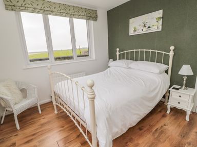 A bedroom with a white metal bed frame white bedding a nightstand with a lamp a wicker chair and a window with a green valance at Seaview Howick in Craster