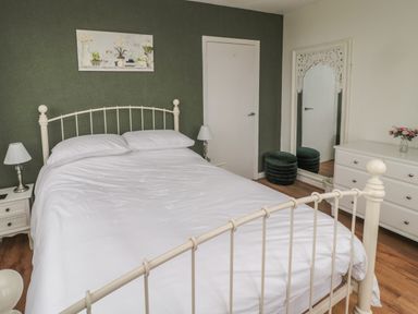 A bedroom with a white bed and bedding two bedside tables with lamps a dresser with flowers a large decorative mirror and a green accent wall at Seaview Howick in Craster