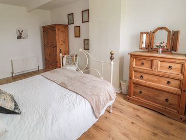 A bedroom with a white metal bed frame a wooden wardrobe a wooden dresser with a mirror and flowers a chair and framed pictures on the wall at Seaview Howick in Craster