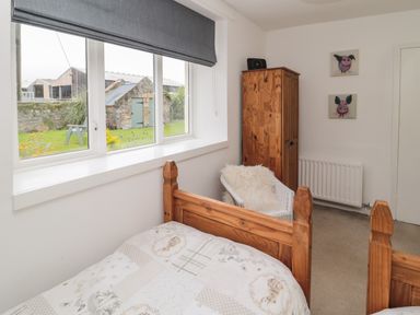 A bedroom with a wooden bed, a white wicker chair with a cushion, a wooden wardrobe, and two pig paintings on the wall at Seaview Howick in Craster