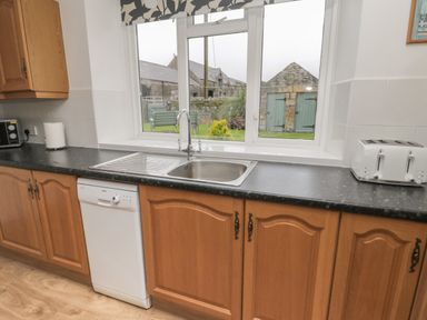 A kitchen with wooden cabinets a sink a dishwasher a toaster and a window overlooking a garden at South Cottage Howick Craster