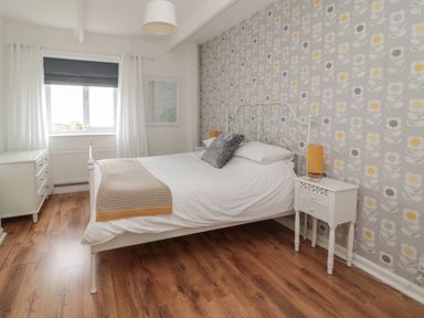 A bedroom with a white bed and patterned wallpaper a bedside table with a lamp and a window with white curtains at South Cottage Howick in Craster