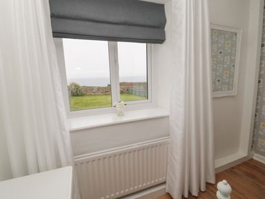 A window with white curtains and gray blind overlooking a garden with a stone wall and sea at South Cottage Howick in Craster