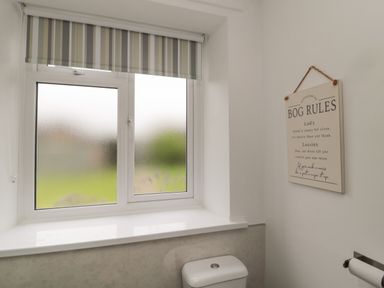 A bathroom window with frosted glass and a striped roller blind above a toilet and a wall sign listing bathroom rules at South Cottage Howick in Craster