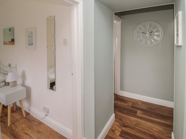 A hallway with a clock on the wall and a bedroom corner with a bedside table lamp and wall art at South Cottage Howick in Craster