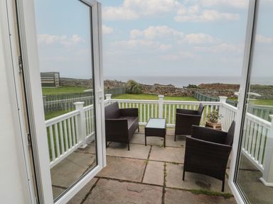 A patio with wicker furniture and a glass table overlooking a grassy yard and stone wall at South Cottage Howick in Craster