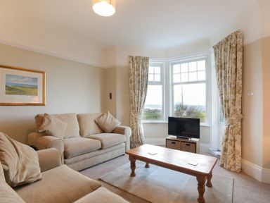A living room with beige sofas a wooden coffee table a television and windows with floral curtains at Ty y Mor in Seahouses