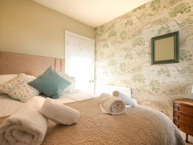 A bedroom with a bed with cushions and rolled towels on a quilt a wooden nightstand and a framed mirror on wallpapered wall at Ty y Mor in Seahouses