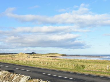 A coastal road with grassy dunes and a sandy beach with the sea in the background at Ty y Mor in Seahouses