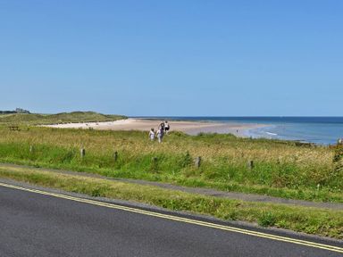 A beach with people walking on a path near grass and a road with double yellow lines at Ty y Mor in Seahouses
