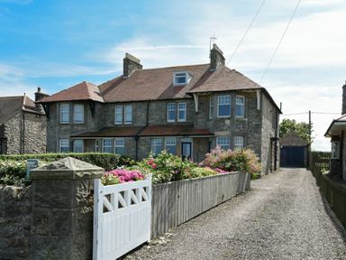 A stone house with multiple windows and a tiled roof next to a gravel driveway and garden with flowers at Ty y Mor in Seahouses