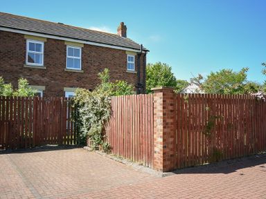 A brick house with two white-framed windows behind a red wooden fence and a paved driveway at Sugar Sands in Longhoughton