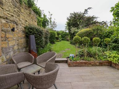 An outdoor patio with wicker chairs and a glass table next to a garden with green bushes and trees at The Book House in Warkworth
