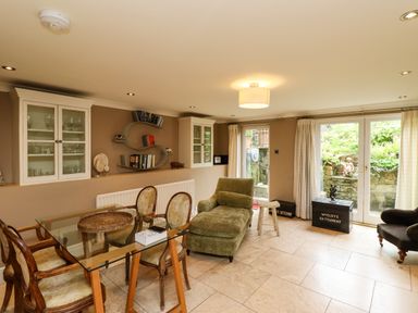 A dining area with a glass table and four chairs a green chaise lounge shelves with books and cabinets and doors leading outside at The Book House in Warkworth
