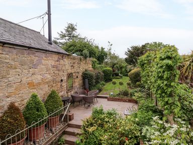 A stone wall with patio furniture and potted plants next to a green garden with trimmed bushes at The Book House in Warkworth