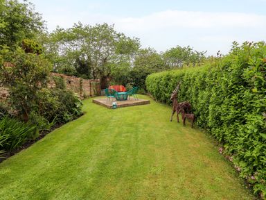 A garden with trimmed grass and hedges a seating area with colorful chairs and a table and two wicker animal sculptures at The Book House in Warkworth
