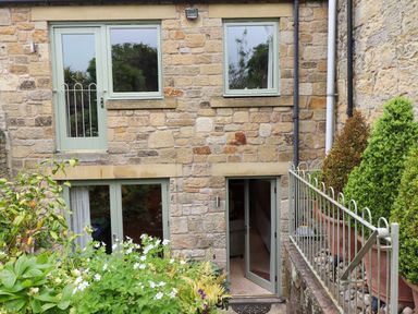 Stone exterior wall of a house with green doors and windows surrounded by plants and a metal railing at The Book House in Warkworth