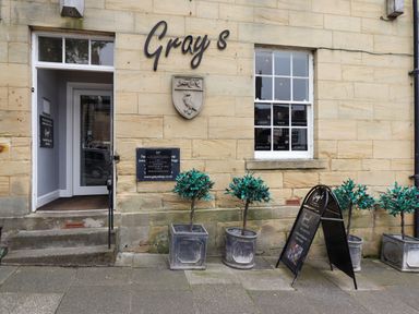 The exterior of a shop with a door, window, potted plants, and signage with the name Grays