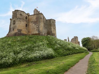 An ancient stone castle on a grassy hill next to a paved path at The Book House in Warkworth