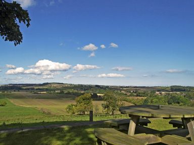 A wooden picnic table on grass with a view of fields and hills under a blue sky at The Huffy House in Alnmouth