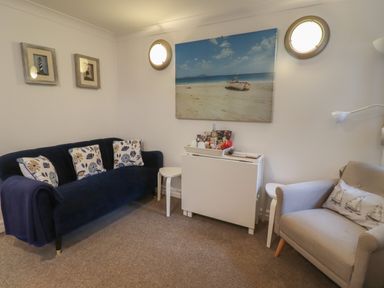 A sitting room with a blue sofa with patterned cushions a beige armchair with a sailboat cushion two small side tables a white cabinet with snacks on top and wall art of a beach and lighthouses at The Little Boathouse in Seahouses