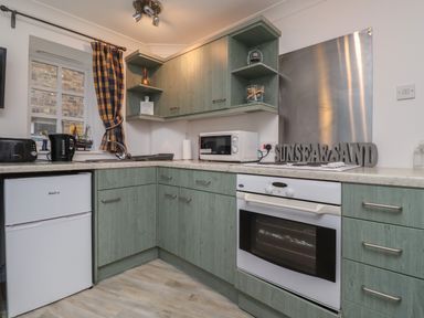 A kitchen with green cabinets a white oven a microwave kettles and a window with a plaid curtain at The Little Boathouse in Seahouses