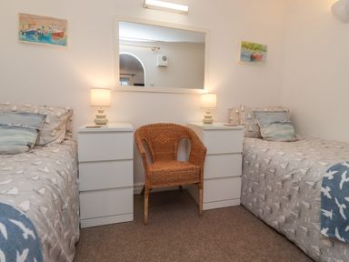 A bedroom with two single beds a wicker chair between two white bedside tables with lamps and a large mirror on the wall at The Little Boathouse in Seahouses