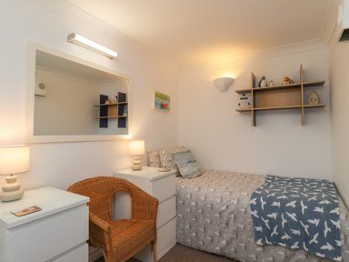 A small bedroom with a single bed a wicker chair two white dressers with lamps and wall shelves at The Little Boathouse in Seahouses