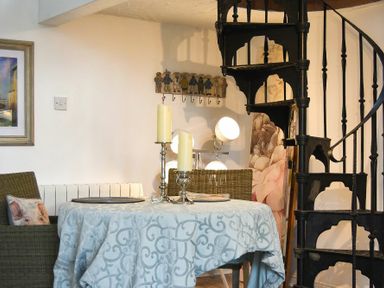 A dining area with a round table covered with a blue patterned tablecloth surrounded by wicker chairs and a black spiral staircase in the corner at The Snug in Embleton