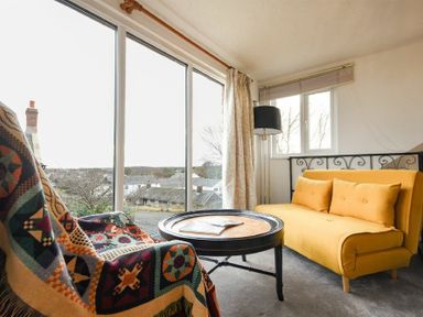 A sitting area with a yellow sofa a patterned blanket on a chair and a round table by large windows at The Snug in Embleton
