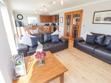 A living area with two black leather sofas and cushions a wooden coffee table with a bowl of decorative items and a lantern a kitchen and dining area with wooden cabinets a table and chairs and a wall clock at Tree Corner in Beadnell