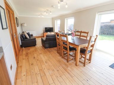 A dining table with six chairs and a vase with flowers next to two leather sofas and a television in a room with wooden floor and windows at Tree Corner in Beadnell