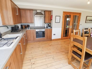 A kitchen with wooden cabinets a countertop a sink a toaster a microwave an oven wooden floor and a dining table with chairs at Tree Corner in Beadnell