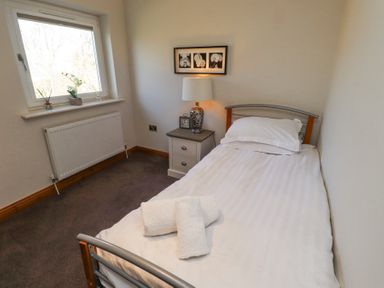 A small bedroom with a single bed white linens a bedside table with a lamp and clock and a window with decor at Tree Corner in Beadnell