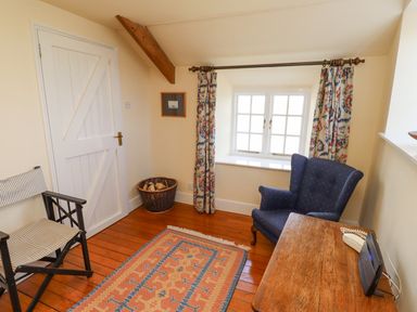 A small room with a wooden floor a patterned rug a blue armchair a wooden table a striped chair a basket of firewood a white door and a window with patterned curtains at Wynding Down in Bamburgh