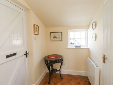 A small room with a white wooden door a round ornate dark wooden table with a book on it a window with a decorative box on the sill framed pictures on cream walls and a white radiator on the floor with terracotta tiles at Wynding Down in Bamburgh