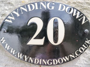 A black oval house sign with white text showing number 20 and website address on a stone wall at Wynding Down in Bamburgh
