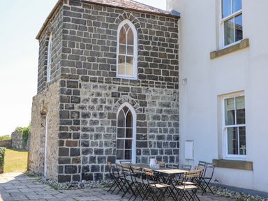 An outdoor patio area with a long wooden table and folding chairs next to a stone building with arched windows at The Lookout Embleton