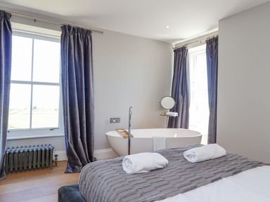 A bedroom with a bed and rolled towels next to a freestanding bathtub and windows with curtains at The Lookout in Embleton