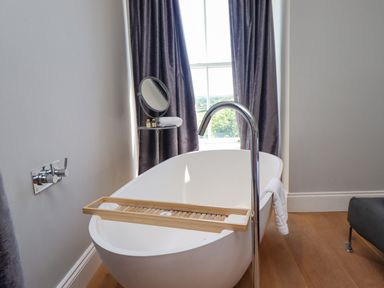 A freestanding bathtub with a wooden bath tray and towel in a bathroom with a window and curtains at The Lookout Embleton