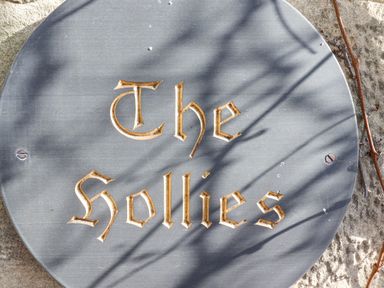 A round stone sign engraved with the words The Sollies mounted on a textured stone wall with shadows and a vine at The Hollies in Bamburgh