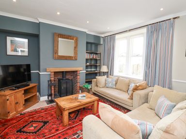 A living room with beige sofas a wooden coffee table a patterned red rug a television on a wooden stand a fireplace and striped curtains at The Hollies in Bamburgh