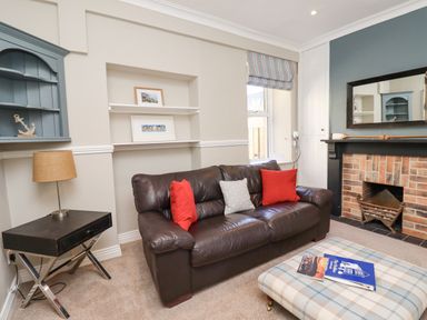 A living room with a brown leather sofa with red and gray cushions a plaid ottoman a fireplace and a side table with a lamp at The Hollies in Bamburgh