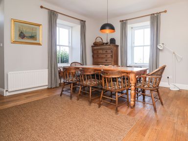 A dining room with wooden table and chairs two windows with curtains a floor lamp and a wooden cabinet at No6 Sea Lane in Embleton