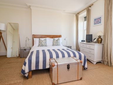 A bedroom with a wooden bed, striped blanket, metal trunk, white dresser with lamp and TV, tall mirror and curtains at No6 Sea Lane Embleton