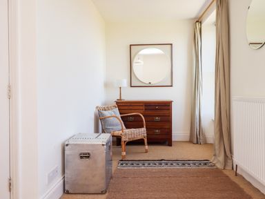 A corner of a room with a wicker chair and cushion a wooden chest of drawers with a round mirror and lamp beige curtains and a metal trunk at No6 Sea Lane in Embleton