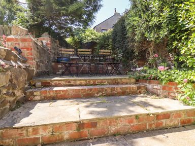 An outdoor seating area with metal chairs and a table on brick steps surrounded by plants at No6 Sea Lane in Embleton