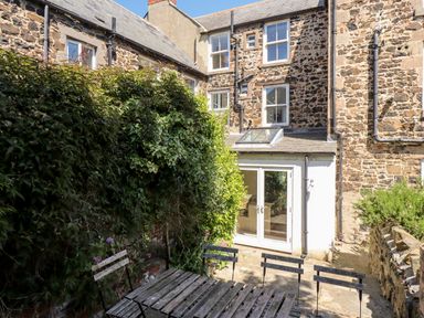 A stone building with multiple windows a patio area with a wooden table and chairs and greenery at No6 Sea Lane in Embleton