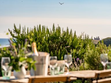 An outdoor table with wine glasses and a bucket with a bottle with greenery and a distant castle by the sea at No6 Sea Lane in Embleton